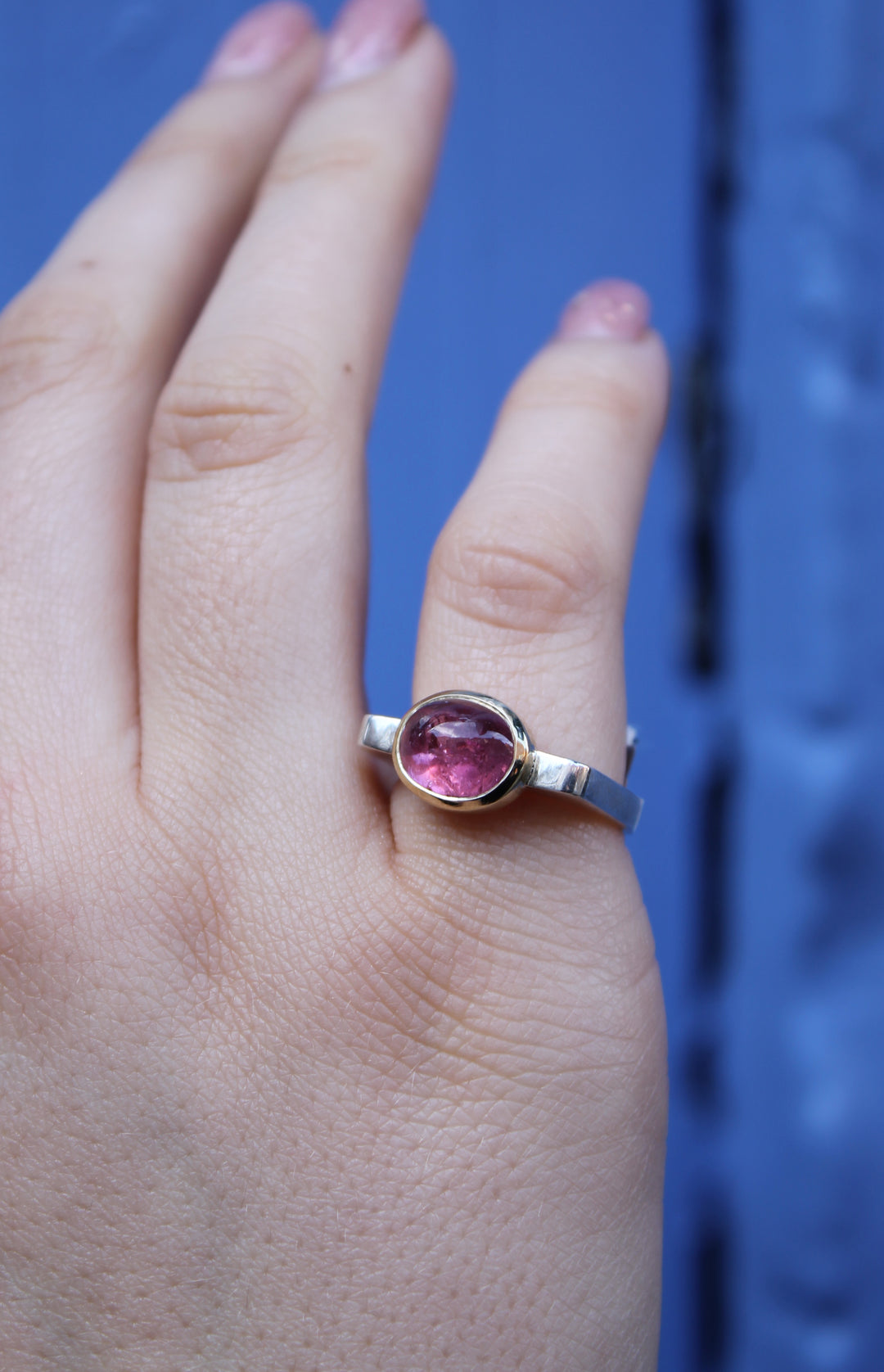 A hand with light pink nail polish showcases the Madeleine Stellar Blush Silver & Gold Ring, featuring an elegant oval cabochon design, set with a pink gemstone, against a blurred blue background.