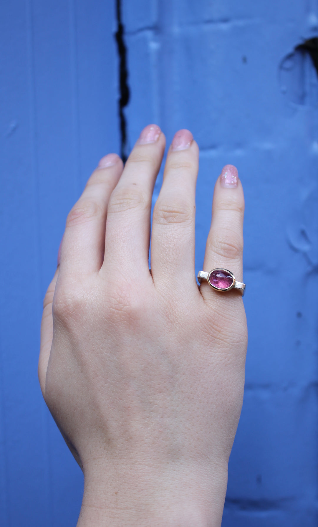 A hand with light pink, glittery nail polish wears the Madeleine Stellar Blush Silver & Gold Ring, featuring a pink gemstone, against a textured blue background.