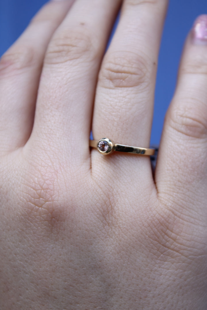 A hand wears the Madeleine Gilded Blossom 9ct yellow gold ring, featuring a small round gemstone at its center, set against a blurred blue background.