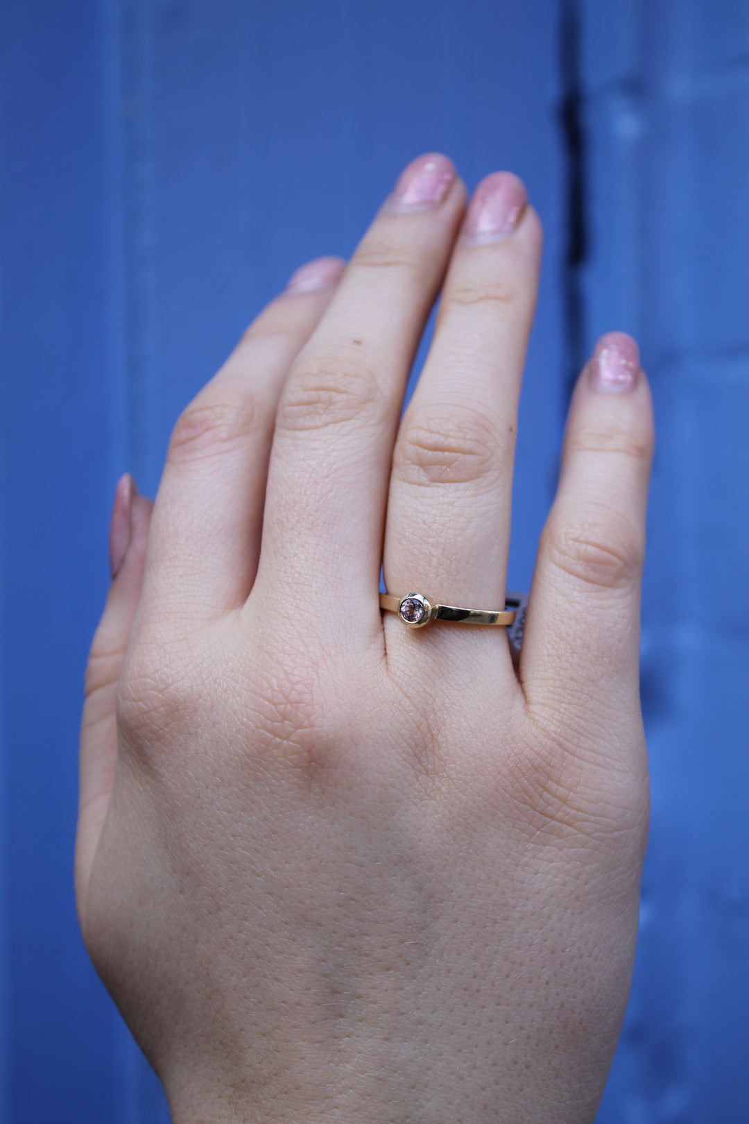 A hand with light pink nails wears the Madeleine Gilded Blossom 9ct yellow gold ring, featuring a delicate cherry blossom design and a small clear stone, set against a blue background.
