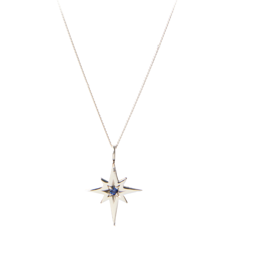 A delicate Polaris Sapphire & Sterling Silver Pendant by Cosmic Boulevard with a celestial pendant in the shape of an eight-pointed star. This handmade jewellery piece features a small, central blue gemstone. The fine chain allows the striking pendant to stand out against the plain white background.