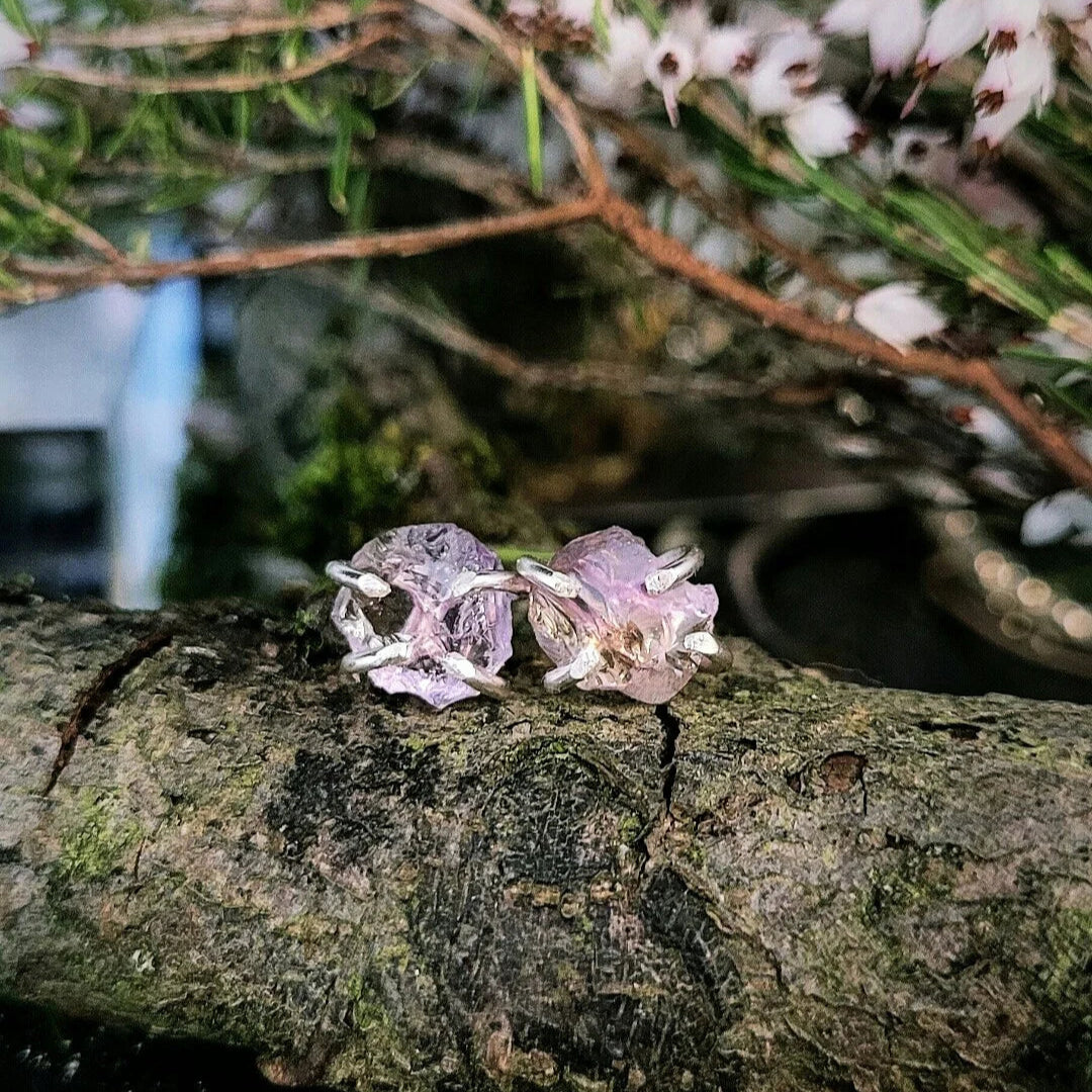 The Raw Ametrine Stud Earrings by Caroline Stokesberry-lee, featuring delicate, light purple flower shapes, rest on a tree branch with blurred greenery and white blossoms in the background.