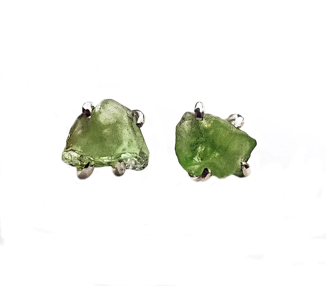 A pair of Raw Peridot Studs by Caroline Stokesberry-Lee featuring raw green peridot gemstones set in sterling silver prongs. The rough, unpolished stones exhibit a natural texture and irregular shape, contrasting with the sleek metal setting. The image has a white background.