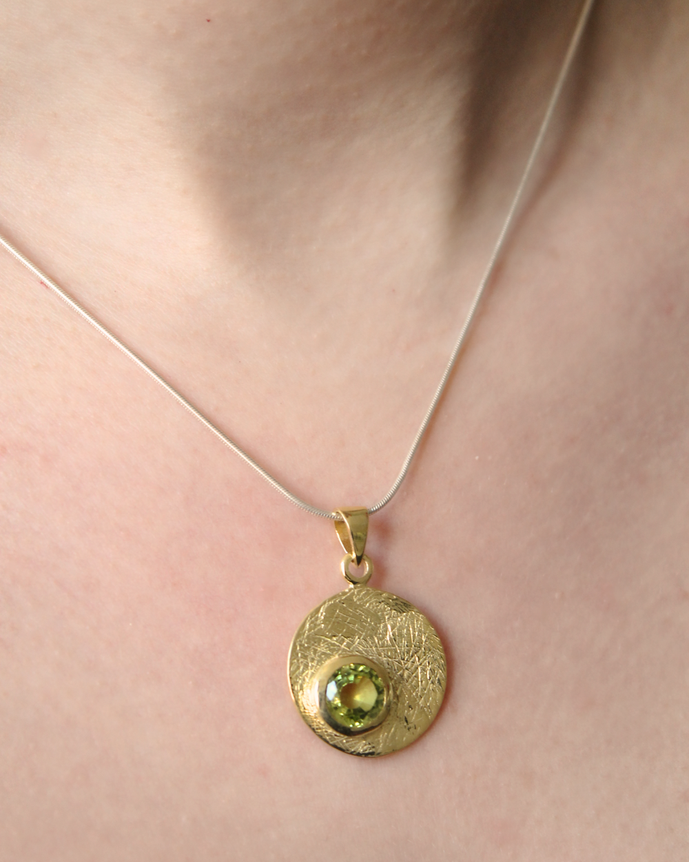 A close-up of a person's neck wearing the Gallardo & Blaine Designs Selene Necklace Gold - Small, featuring a thin chain with a round, textured gold pendant set with a green gemstone.