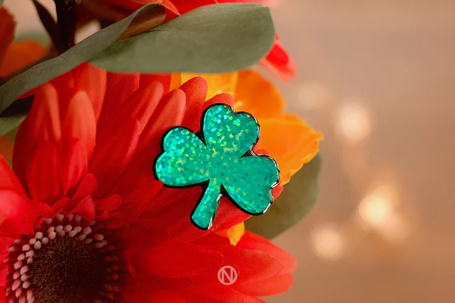 The Naoi Shamrock Pin Brooch sparkles in green atop vibrant orange and red artificial flowers with lush green leaves, softly illuminated by warm, blurred background lights.