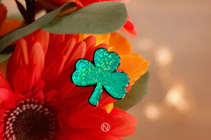 The Naoi Shamrock Pin Brooch sparkles in green atop vibrant orange and red artificial flowers with lush green leaves, softly illuminated by warm, blurred background lights.