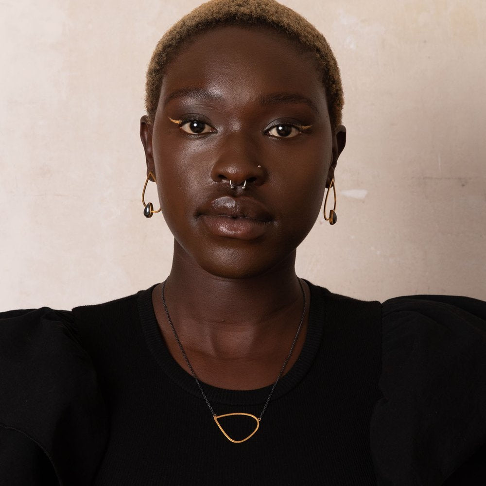 A person with short blonde hair, wearing black and gold earrings, a Leoma Drew Gold Wing Pendant with Oxidised Silver Chain necklace, and a black top with puffed sleeves. They have gold eyeliner and a septum piercing, standing against a neutral textured background.