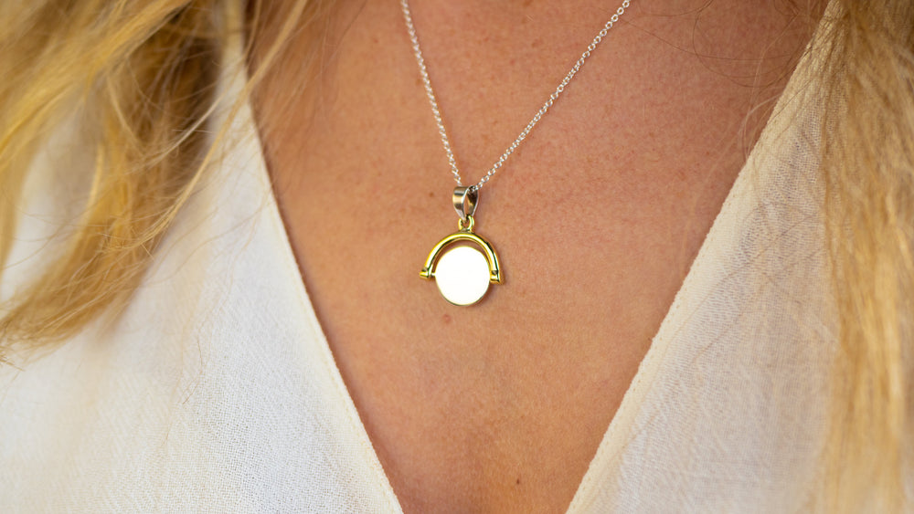 A close-up of a person wearing Engravable Jewellery's Gold Plate Spinning Disc necklace, a customizable piece featuring a gold framed pendant, resting against a light beige V-neck top.