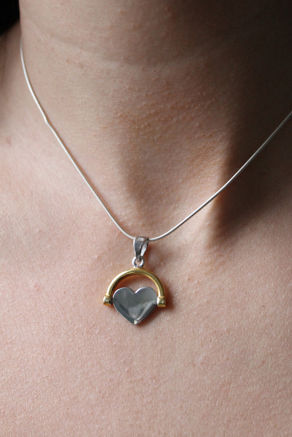 A close-up shows a person's neck wearing Engravable Jewellery's Gold Plate Spinning Heart necklace—a gold plated silver piece with a gray heart framed by a gold arch. Personalize it with custom engraving for a unique touch.