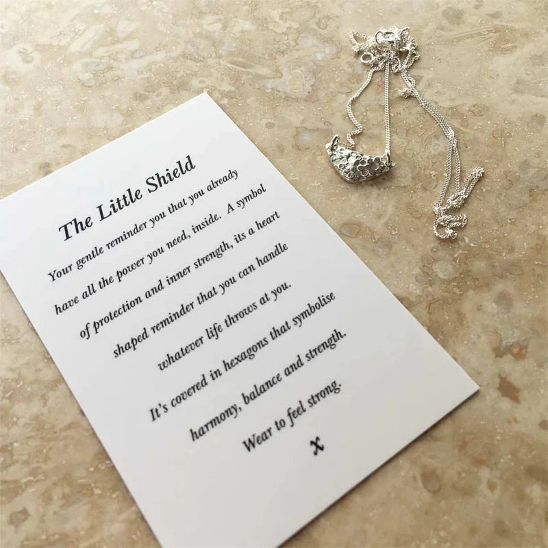 A card with an inspiring message rests on a light stone surface beside the Andrea Mears Jewellery Little Shield Necklace Silver, featuring a sterling silver hexagon-patterned pendant—an elegant touch for everyday wear.