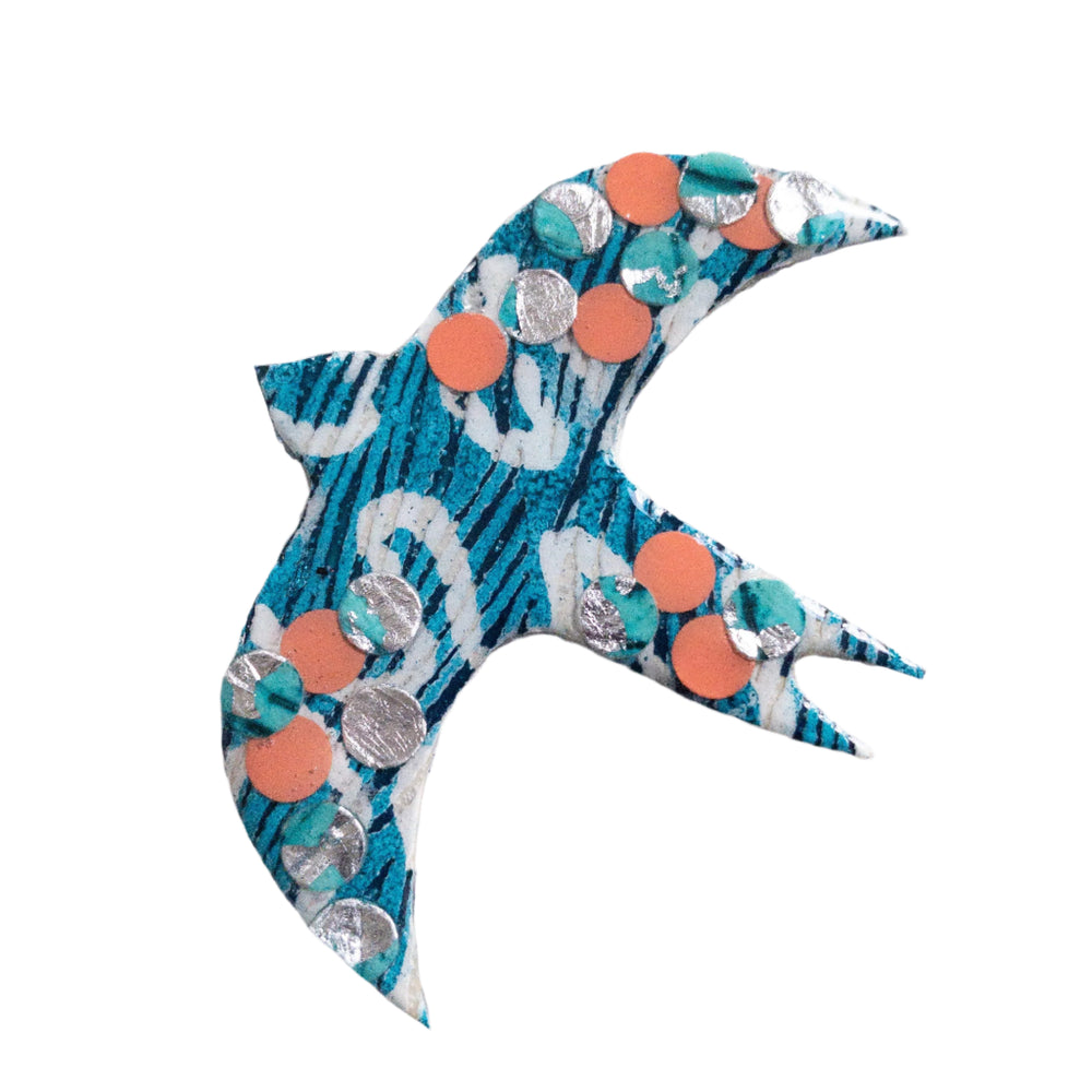 A handmade Éan Bird Batik Textile Brooch by Rothlú with a blue and white pattern, adorned with small orange and silver circular sequins. The bird's wings and tail are spread outward, creating a sense of motion. Its unique texture is set against a plain white background, highlighting the eco resin finish.
