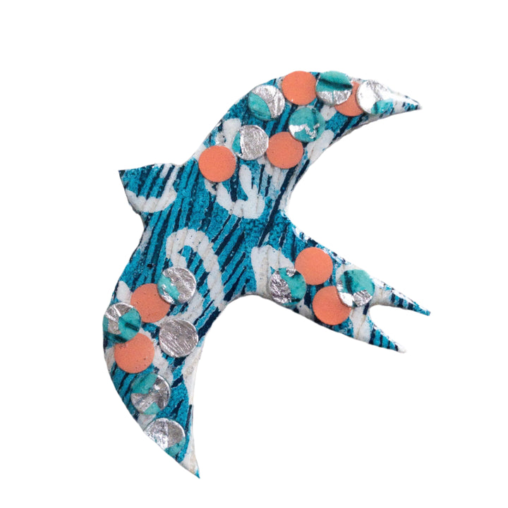 A handmade Éan Bird Batik Textile Brooch by Rothlú with a blue and white pattern, adorned with small orange and silver circular sequins. The bird's wings and tail are spread outward, creating a sense of motion. Its unique texture is set against a plain white background, highlighting the eco resin finish.