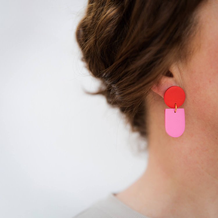 Close-up of a woman's ear wearing Tengae Red & Pink Earrings by Culu Design—handmade dangles with a red circular top and pink rectangular drop, styled with her brown hair up against a softly blurred background.