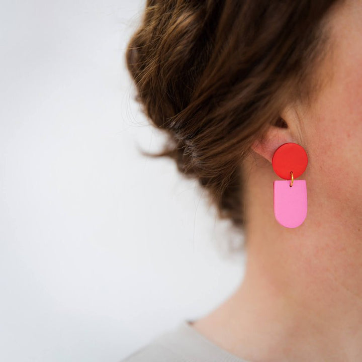 Close-up of a woman's ear wearing Tengae Red & Pink Earrings by Culu Design—handmade dangles with a red circular top and pink rectangular drop, styled with her brown hair up against a softly blurred background.