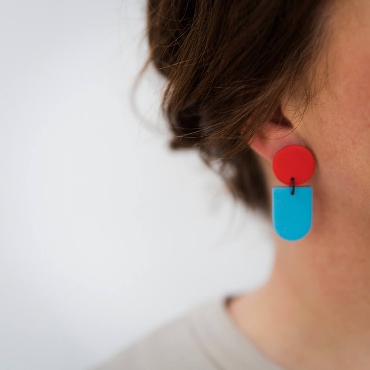 Close-up of a person wearing Culu Design's Tengae Red & Robin's Egg Earrings—handmade polymer clay dangles with a red circular top and blue semi-oval bottom. The person's brown hair partly covers their ear against a white background.