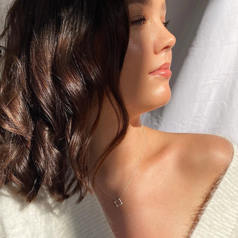 A woman with wavy brown hair and radiant skin wears a white top and the Waves Necklace Gold by Andrea Mears Jewellery—an 18ct gold plated piece with a small open square pendant. Sunlight and a white curtain frame this ocean-inspired accessory.
