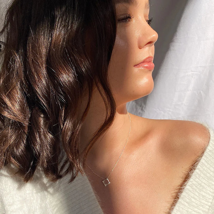 A woman with wavy brown hair and radiant skin wears a white top and the Waves Necklace Gold by Andrea Mears Jewellery—an 18ct gold plated piece with a small open square pendant. Sunlight and a white curtain frame this ocean-inspired accessory.