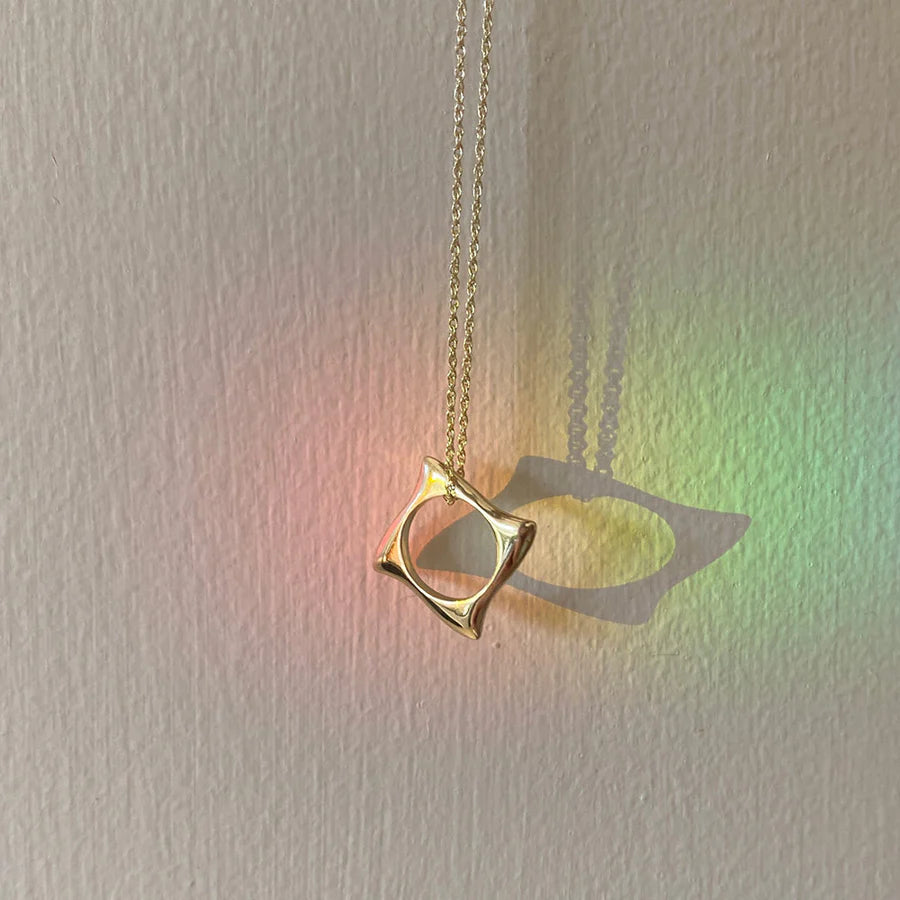 The Waves Necklace Gold by Andrea Mears Jewellery features a gold wave pendant in an abstract square frame, casting soft shadows on a beige wall with subtle pastel rainbow reflections—an ideal piece of ocean-inspired jewelry.