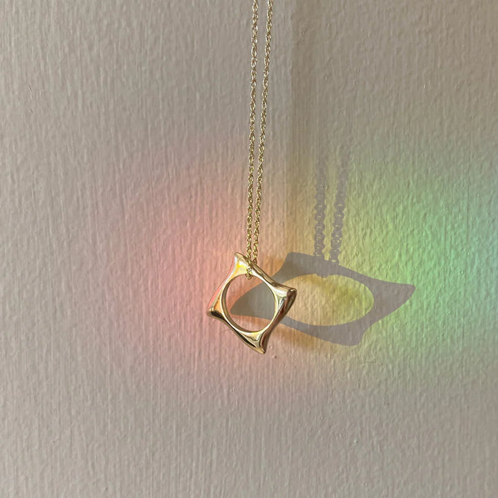 The Waves Necklace Gold by Andrea Mears Jewellery features a gold wave pendant in an abstract square frame, casting soft shadows on a beige wall with subtle pastel rainbow reflections—an ideal piece of ocean-inspired jewelry.