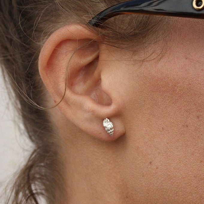 A close-up shot of a person's ear wearing Jennifer Kinnear's Whelk Shell Stud Earrings. The person is also wearing black-framed glasses. Their ear and a portion of their brown hair are visible in the image, with a light background partially visible.