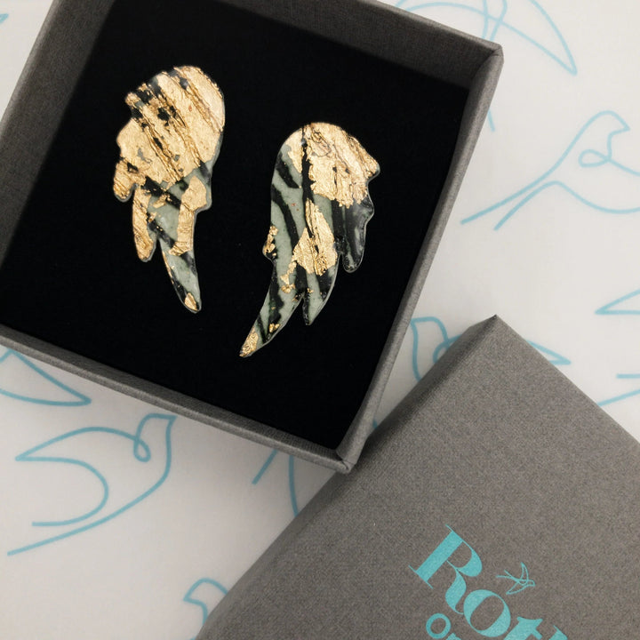 A pair of intricately designed wing-shaped Wings of Desire Sgraffito Stud Earrings with black and gold accents are displayed in a black-lined grey box, which has the word "Rothlú" partially visible. Featuring unique textures and sterling silver posts, the background is decorated with a minimalist bird pattern in light blue.