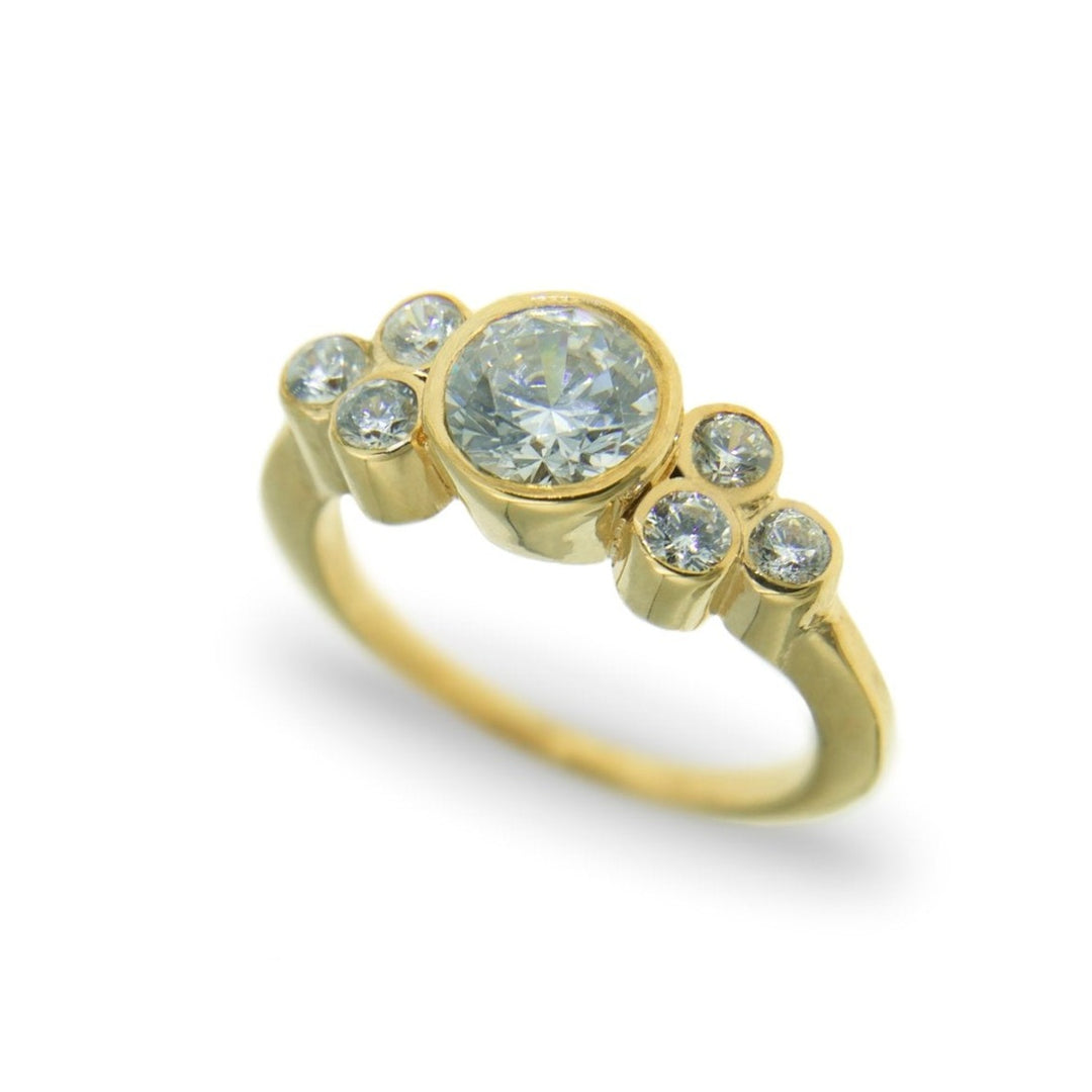 The Miriam Wade Sionna - Seven Stone Engagement Ring features a central 1 carat lab grown diamond flanked by three smaller round diamonds on each side, arranged in a graduated size pattern. The ring has a polished finish, and the diamonds are set in individual gold settings, creating a symmetrical design.