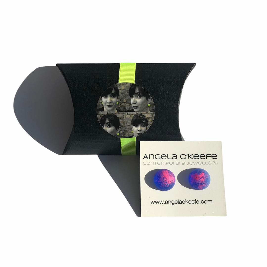 A black, pillow-shaped gift box decorated with a circular photo of a woman’s face, secured with a green ribbon. Next to it is a white card with "Angela O'Keefe" printed on it and featuring bright-colored pink and blue Graffiti Stud earrings crafted in sterling silver.
