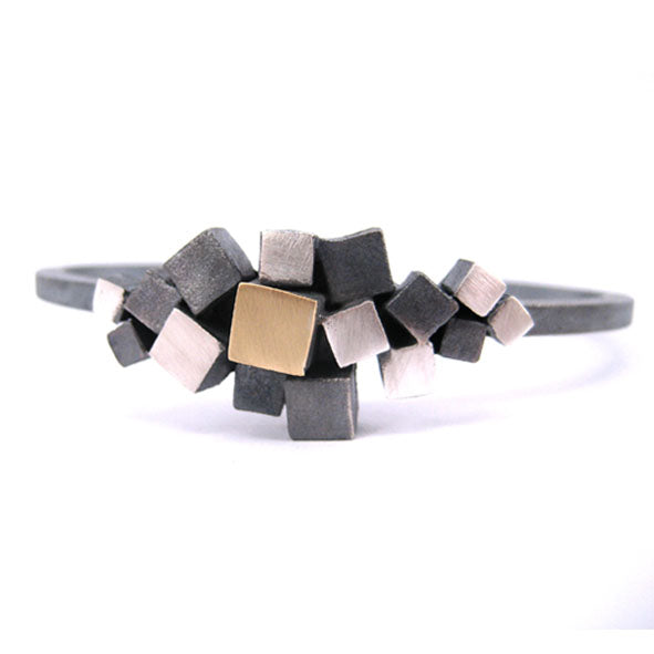 The MarCucurella Iceland Bracelet features a cluster of small cubes in various shades of silver and a central gold cube, arranged asymmetrically on a dark, rigid band. Part of our Iceland collection, this piece captures the raw allure of Reynisfjara beach, creating a modern, abstract look.