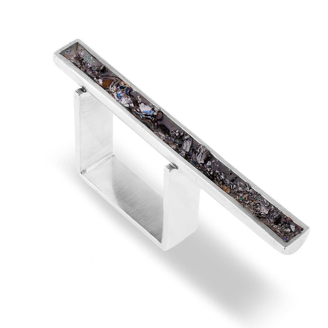 A modern, rectangular silver ring featuring a horizontal bar encrusted with a textured, dark crystalline material. The Angela O'Keefe Long Silver & Black Ring has an open design with a sleek, minimalist aesthetic and subtle grey tones reminiscent of Seville's sophisticated charm.