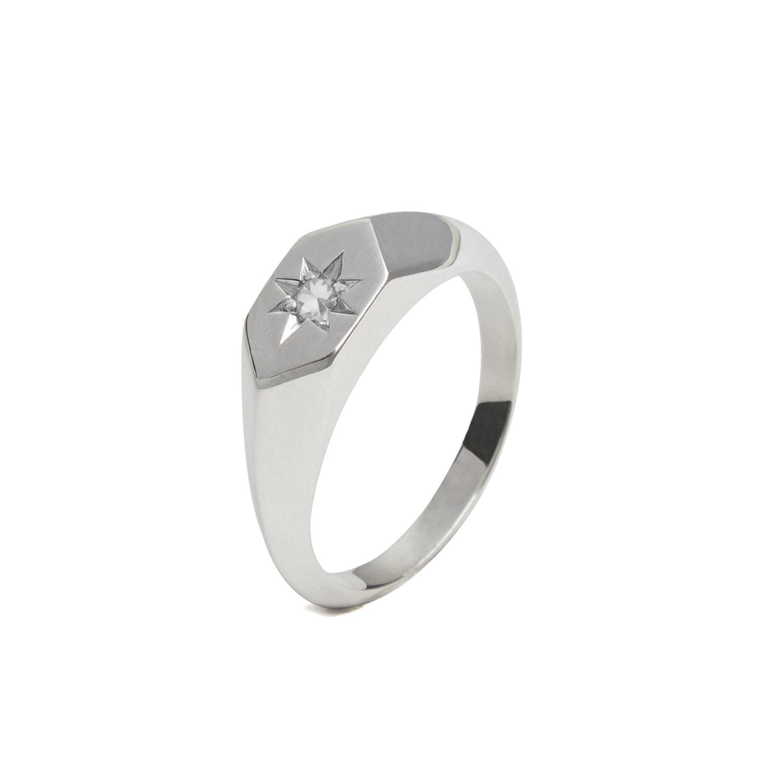 A Starlight Diamond Birthstone Signet Ring from Cosmic Boulevard with a flat hexagonal top displays a small, star-shaped design in the center, embedded with a clear, sparkling gemstone. Handmade in Dublin, this sleek and modern piece features polished and clean lines.