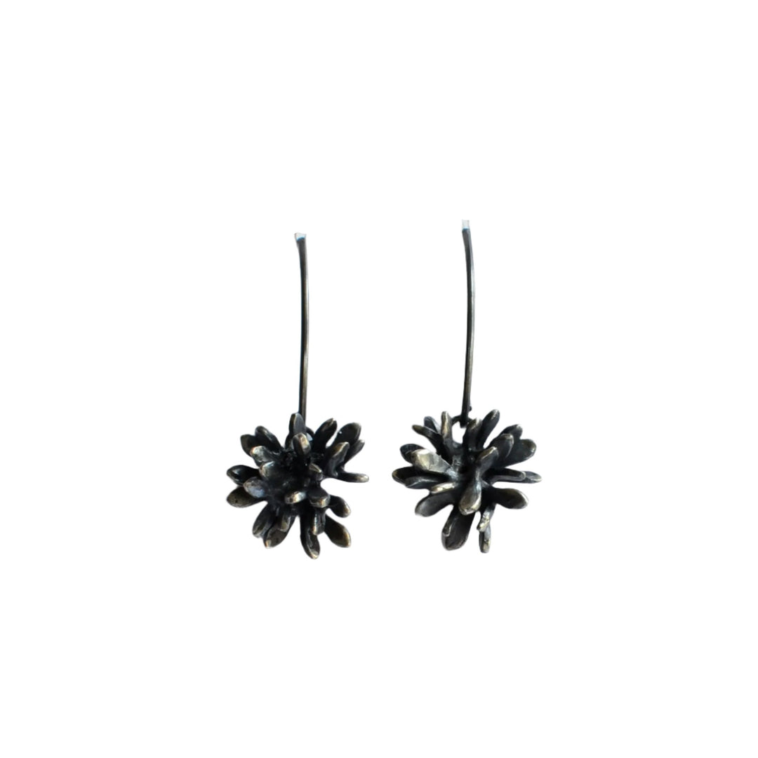 A pair of **Abbie Dixon Folia Oxidised Silver Drop Earrings** featuring intricately designed flower-shaped pendants with delicate leaf shapes. The earrings have long, slender hooks and an oxidised finish that gives the flowers a subtly textured appearance. They are displayed against a plain white background.