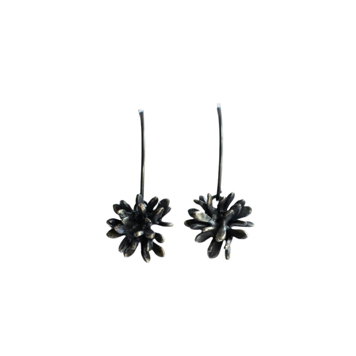 A pair of **Abbie Dixon Folia Oxidised Silver Drop Earrings** featuring intricately designed flower-shaped pendants with delicate leaf shapes. The earrings have long, slender hooks and an oxidised finish that gives the flowers a subtly textured appearance. They are displayed against a plain white background.