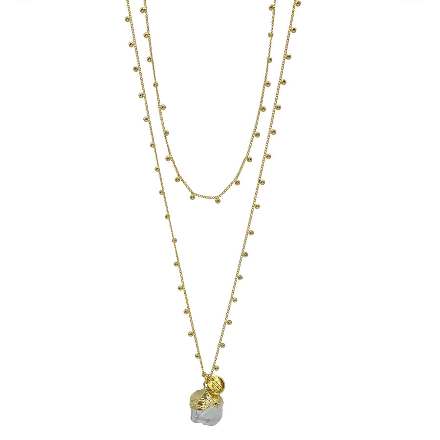 The Khione Necklace by Aria V is a layered piece featuring 16k gold plated chains. One chain is adorned with small, evenly spaced bead-like decorations, while the lower chain holds a large, irregularly shaped clear quartz pendant and a smaller, round, textured charm. The necklace has a delicate and elegant design.