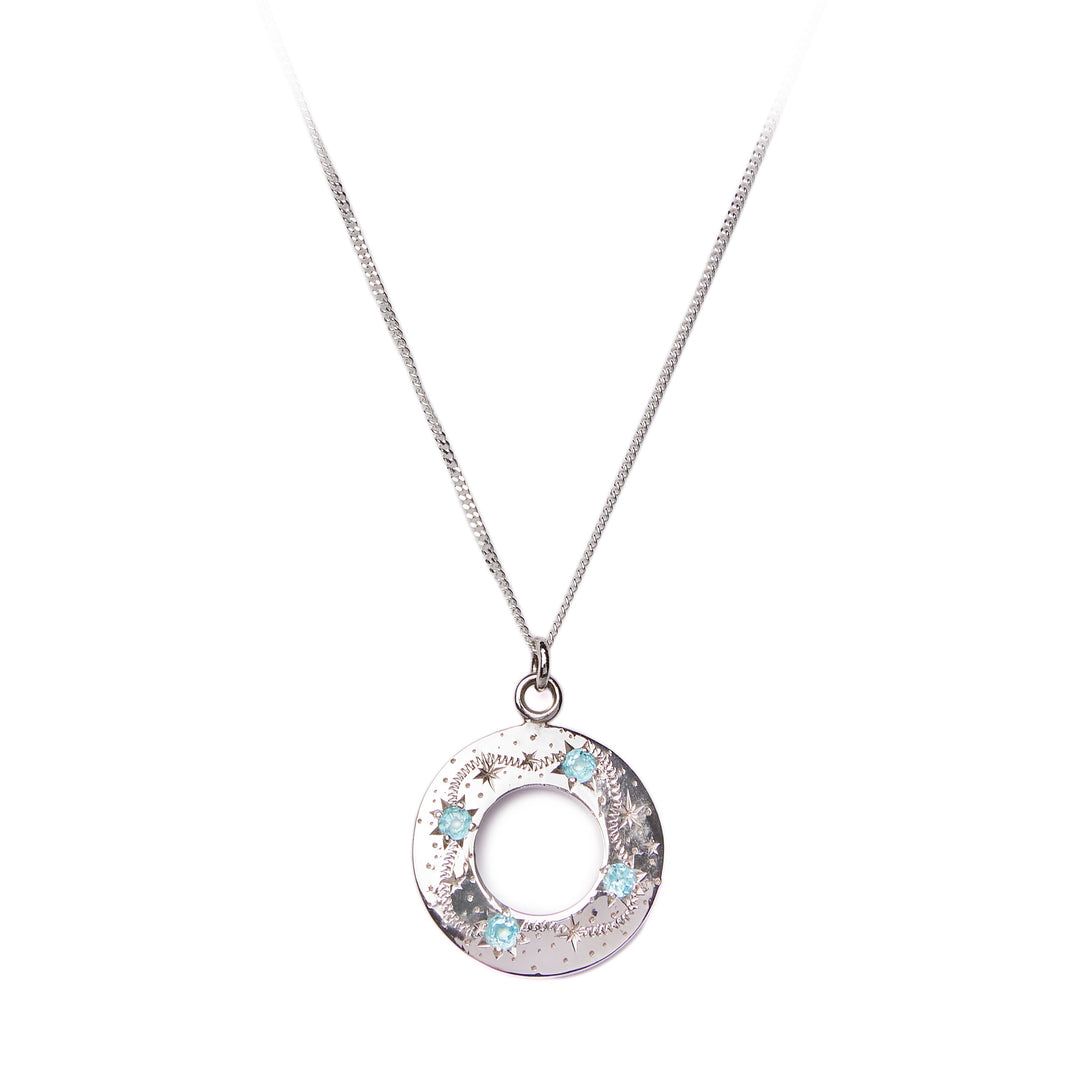 Introducing the "Something Blue" Shooting Star Pendant from Cosmic Boulevard. This exquisite necklace features a circular pendant intricately engraved with patterns and adorned with blue topaz gemstones, reminiscent of shooting stars. The delicate chain enhances the subtle elegance of this silver piece, making it a perfect accessory for sky lovers.