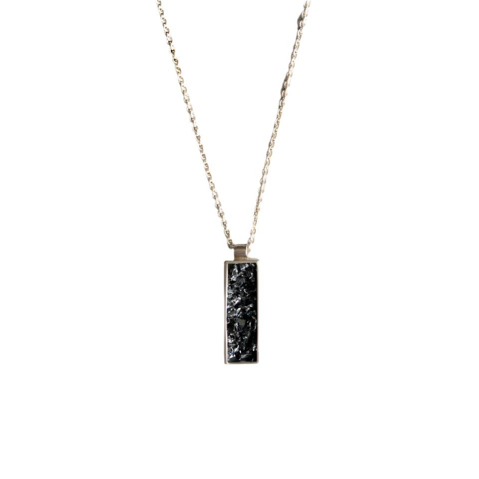 An Ingot Pendant by Angela O'Keefe is shown. The pendant features a rough black stone, possibly crushed silicon carbide, encased in an intricate silver frame. The necklace chain is visible, set against a background composed of contrasting black and light gray areas, evoking a gothic fairy tale aesthetic.