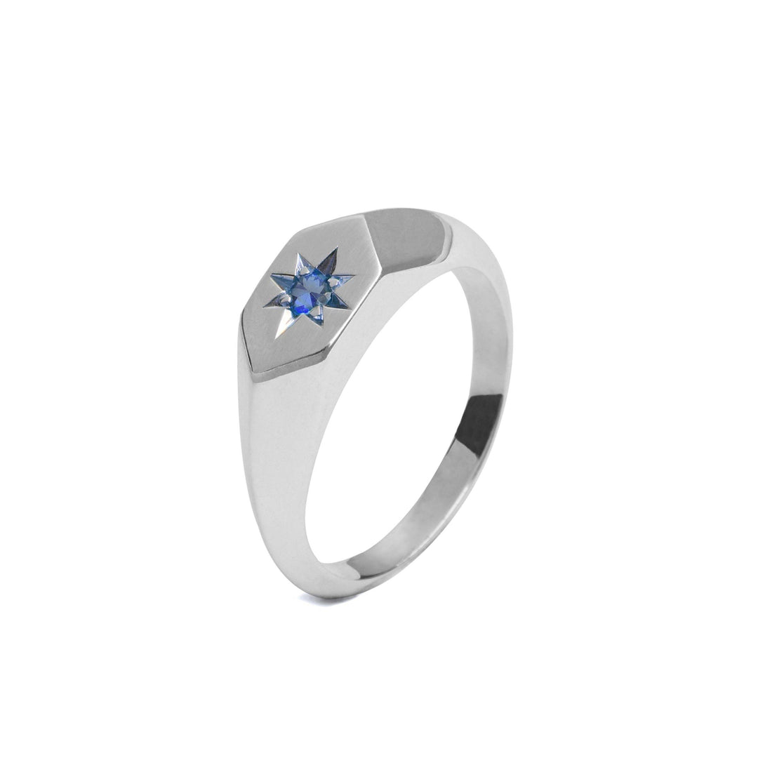 A Cosmic Boulevard Starlight Sapphire Birthstone Signet Ring featuring a star-shaped design on its top, adorned with a small sapphire birthstone at the center of the star. Handmade in Dublin, the ring has a hexagonal embellishment surrounding the star and a polished, smooth band. The background is white.