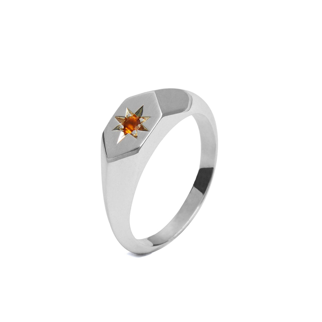 A sleek metallic Starlight Palmeria Citrine Birthstone Signet Ring by Cosmic Boulevard featuring a hexagonal face with a central yellow gemstone set in a star-shaped design. The sterling silver ring’s band is smooth and polished, giving it a modern and elegant appearance. The ring rests against a plain white background.
