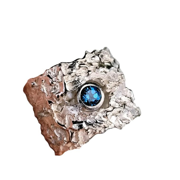 A person holds a uniquely textured "That Place Beyond  Wide Silver Ring with Gemstone" by Caroline Stokesberry-lee, part of the 2022 Collection, adorned with a dark blue gemstone at its center. The wide ring has an irregular, rugged design that gives it an organic appearance. The background is out of focus, emphasizing the intricate details of the ring.