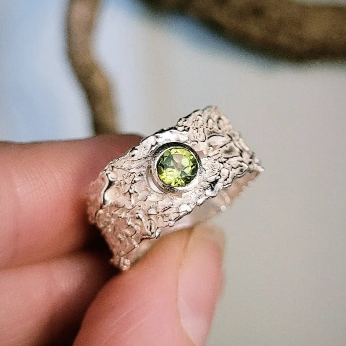 A hand holds the Caroline Stokesberry-lee That Place Beyond Silver Slim Band Ring with a round peridot gemstone at the center, set against a blurred background.
