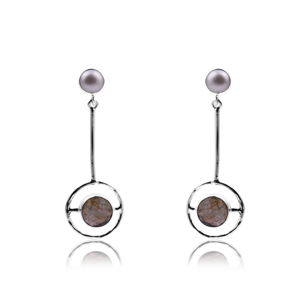 A pair of modern, dangling earrings featuring round, purple pearl studs. The earrings have thin, sterling silver vertical bars with a circular, open-frame charm at the bottom that holds an iridescent labradorite stone in the center. This handmade jewellery design is minimalist and elegant. Introducing the Labradorite and Pearl Pendulum Silver Earrings by Watermelon Tropical.