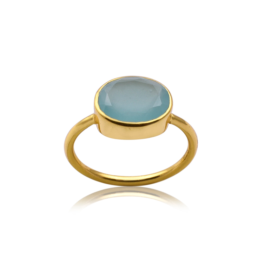 An Oval Aqua Chalcedony Gold Ring by Watermelon Tropical featuring a round, faceted aqua chalcedony gemstone set in a simple bezel setting. This piece of handmade jewellery is displayed on a white background, with a slight reflection visible beneath it.