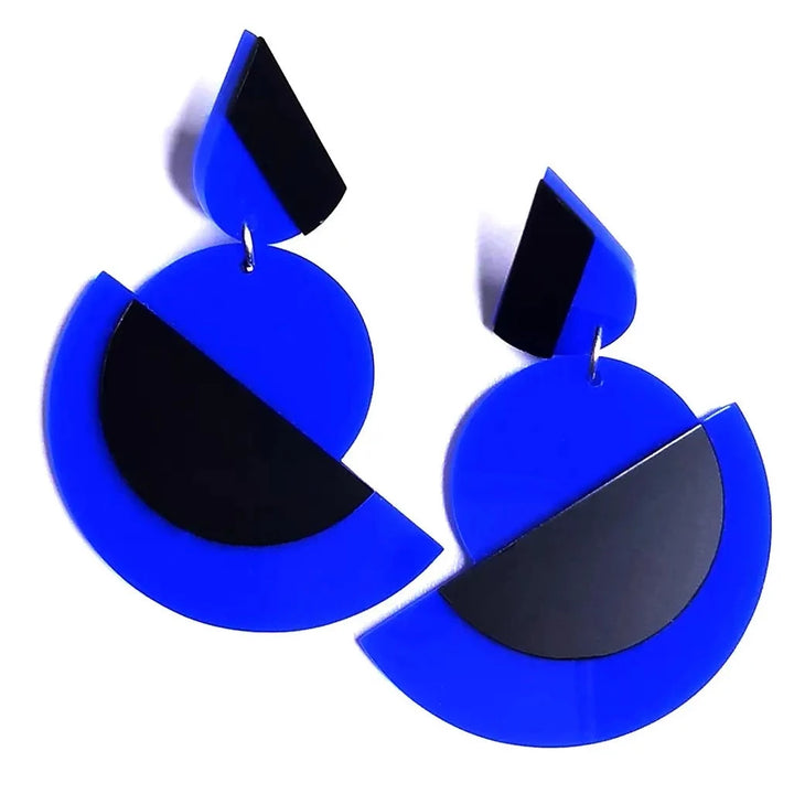 A pair of modern, geometric graphic earrings made of vibrant blue plastic with black accents. Each earring features a circular base and a smaller semi-circular cutout, connected to a black trapezoid at the top—perfect for those who appreciate Avant-Garde aesthetics. Introducing the Suzanna statement acrylic earrings by Alex + Svet.