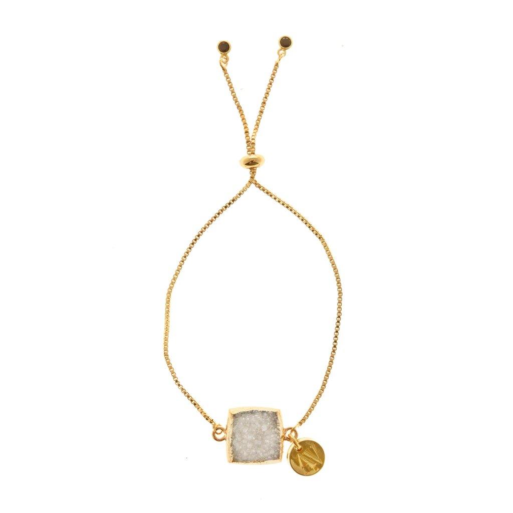 The Aria V Coco Bracelet showcases a gold chain adorned with a square white Druze stone set in a gold frame and a small round gold charm. This elegant piece, beautifully gold plated, features an adjustable slider clasp with two gold beads at the ends of the chain.