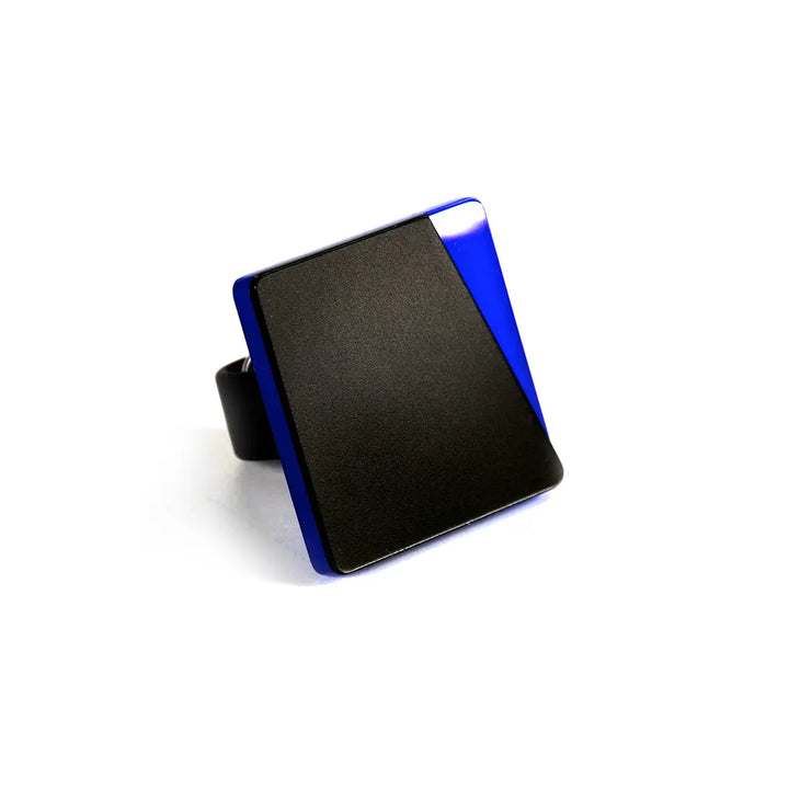 A modern, minimalist ring featuring a large, graphic square design with black and blue geometric color blocks on the top. The band is high-quality acrylic glass, complementing the sleek, angular aesthetic of the ring's surface. The Saylor Ring by Alex + Svet is positioned upright, showcasing its striking design.
