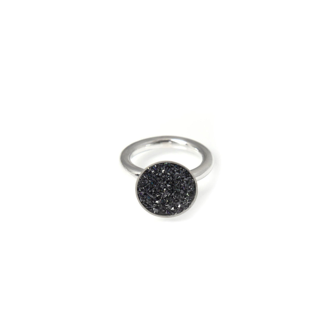 A silver ring featuring a round black-stone inset with a rough, sparkly texture of crushed silicon carbide. The sleek and minimalist band complements the bold, glittering centerpiece. The Angela O'Keefe Black 12mm Cup Ring is displayed against a white background.