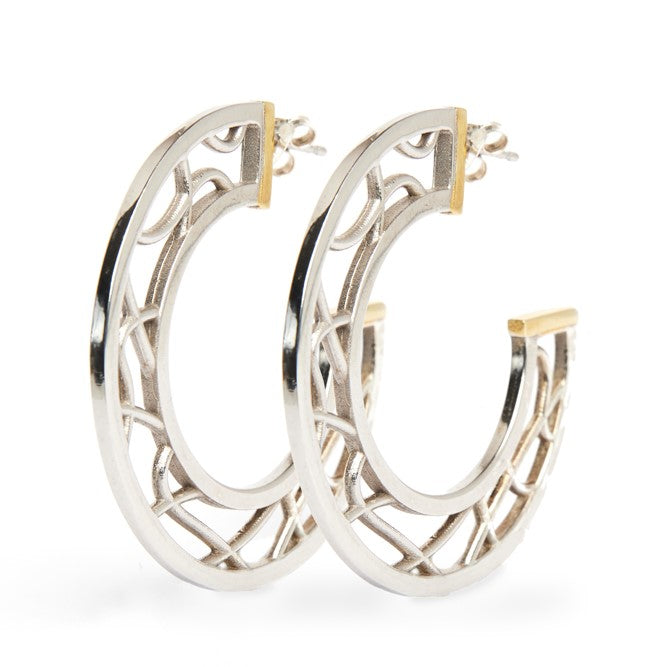 The Maya Earrings by Gallardo & Blaine Designs are a pair of intricately designed sterling silver hoop earrings with gold vermeil accents. The hoops feature open latticework in a geometric pattern, combining modern and classic elements, and are positioned upright to showcase their detailed craftsmanship.