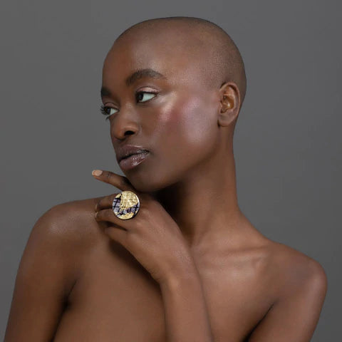 A bald woman against a gray background poses with her head turned to the side and her finger resting on her cheek, showcasing the Ró Cocktail Ring in Gold/Black by Rothlú. She has a serene expression and highlighted cheekbones.