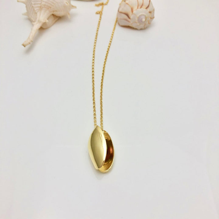 The Open Clam Shell Necklace With Pearl by Jennifer Kinnear, crafted from gold plated silver, is displayed alongside two seashells on a white background. The clam shell-shaped locket boasts a shiny, smooth surface. The intricate shells add a beach-themed aesthetic to the scene.