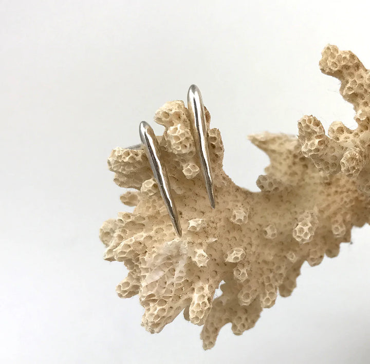 A pair of delicate, sterling silver minimalist earrings displayed against a textured, beige coral background. The Sea Urchin Spine Earrings - Silver by Jennifer Kinnear have a simple, sleek design and are positioned to highlight their smooth, reflective surface.