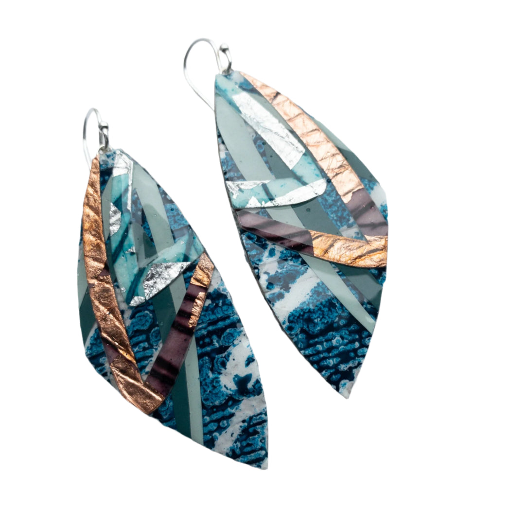 Rothlú’s Leamhan Batik Textile Earrings feature a leaf-inspired design in teal, aqua, silver, rose gold, and aubergine. These handmade earrings boast marbled textures and metallic accents for a unique, artistic touch.